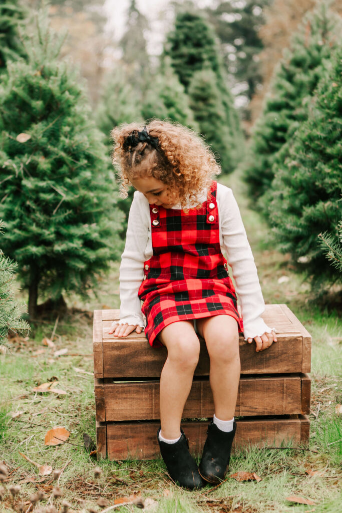 A young girl with curly hair looks down at her hand while sitting on a crate at a Christmas tree farm Skyline Christmas Tree Farm Salem, OR