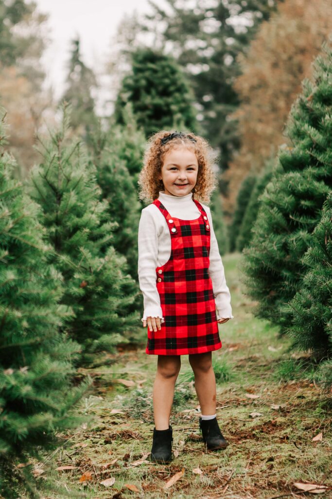 A small girl in a black and red plaid dress smiles while standing in a Christmas Tree farm Salem, OR