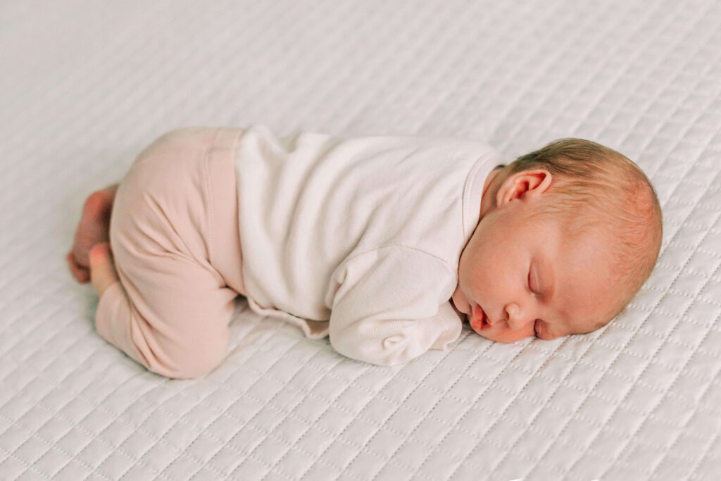 A one month old baby in pink lays on a white comforter