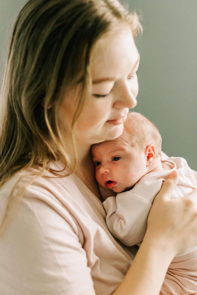 A mother holds her young infant to her chest. They're both wearing soft pink. She has her hand on the baby's back.