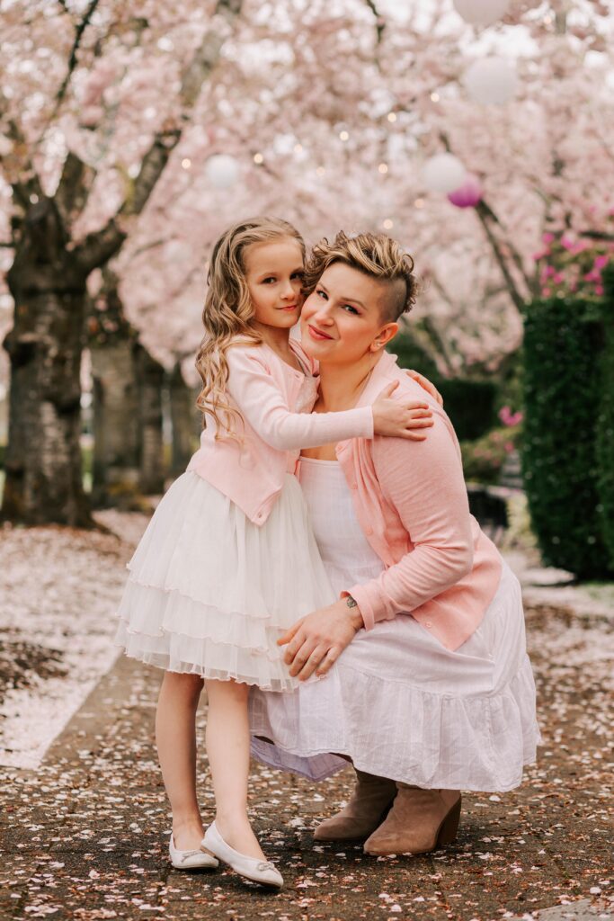 A young girl and her mother in the cherry blossoms in Salem, OR. They are wearin white dresses and pink sweaters and hugging.