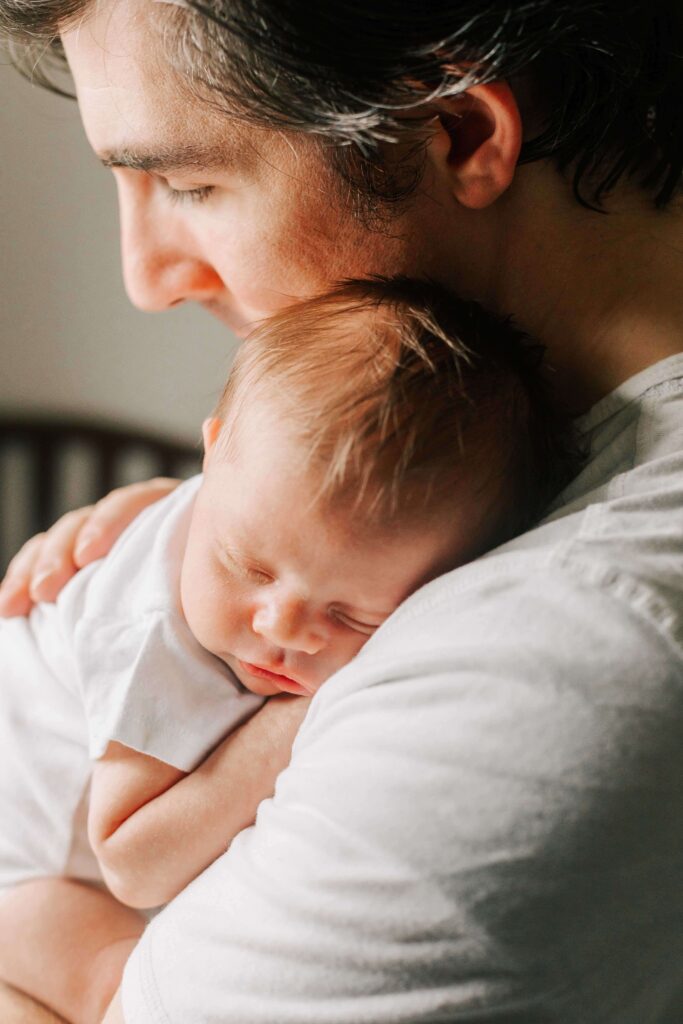 A father cuddles his newborn sleeping baby. Both are in white shirts.