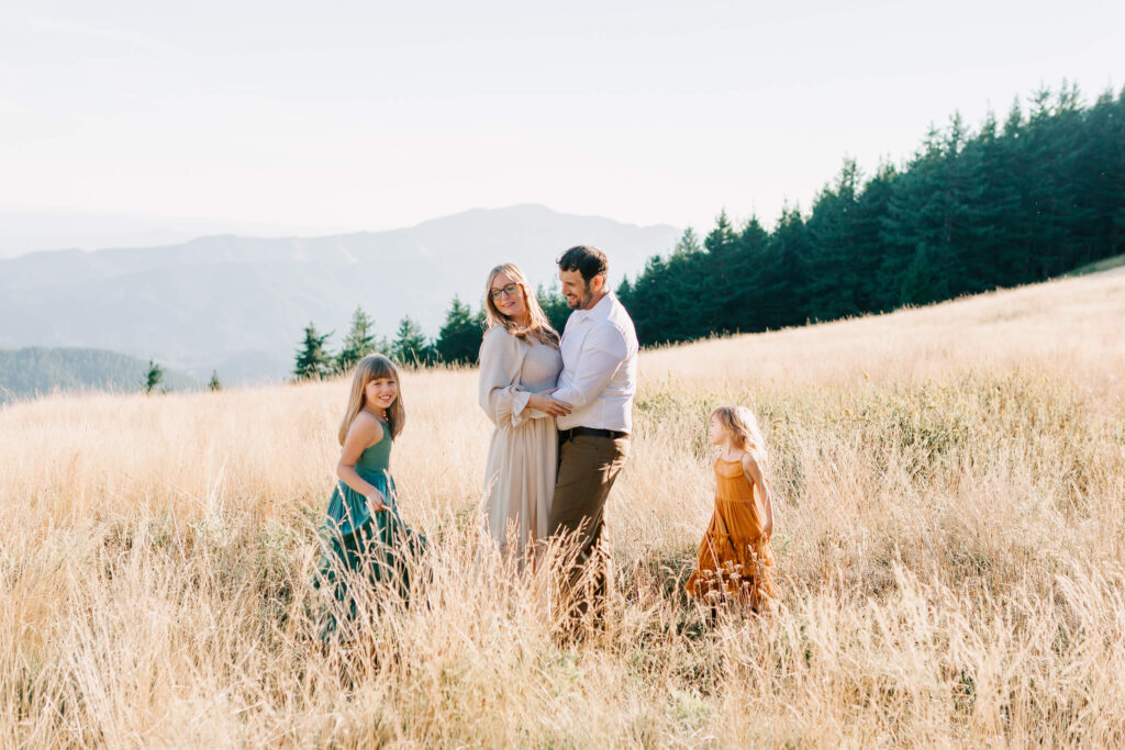 A mother and father hug and watch their children play in a field of tall grass