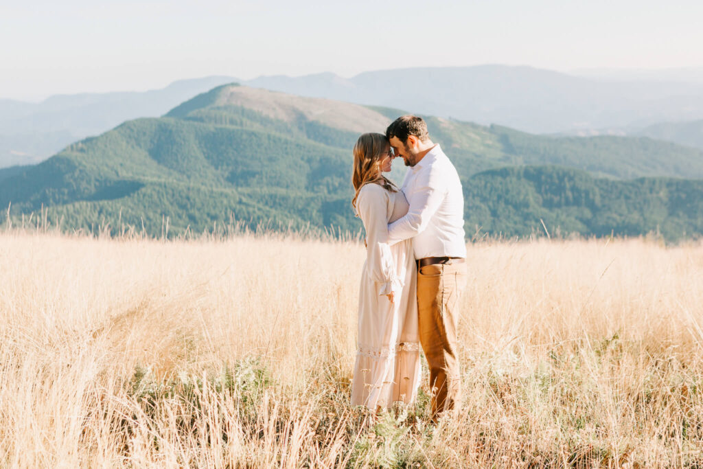 A man and woman stand together looking at each other. They are in a field with mountains in the background. She is in a dress and he is wearing a white shirt and slacks.