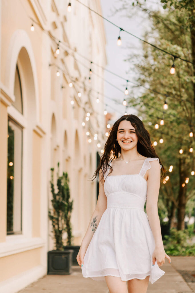 High school senior poses downtown Albany, Or for senior portraits. She's in a white dress with string lights.