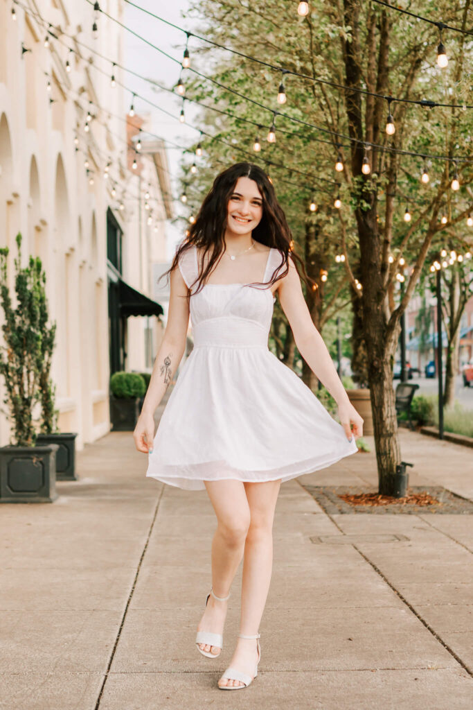 High school senior twirling in a white dress under string lights in downtown Albany, Oregon during golden hour.