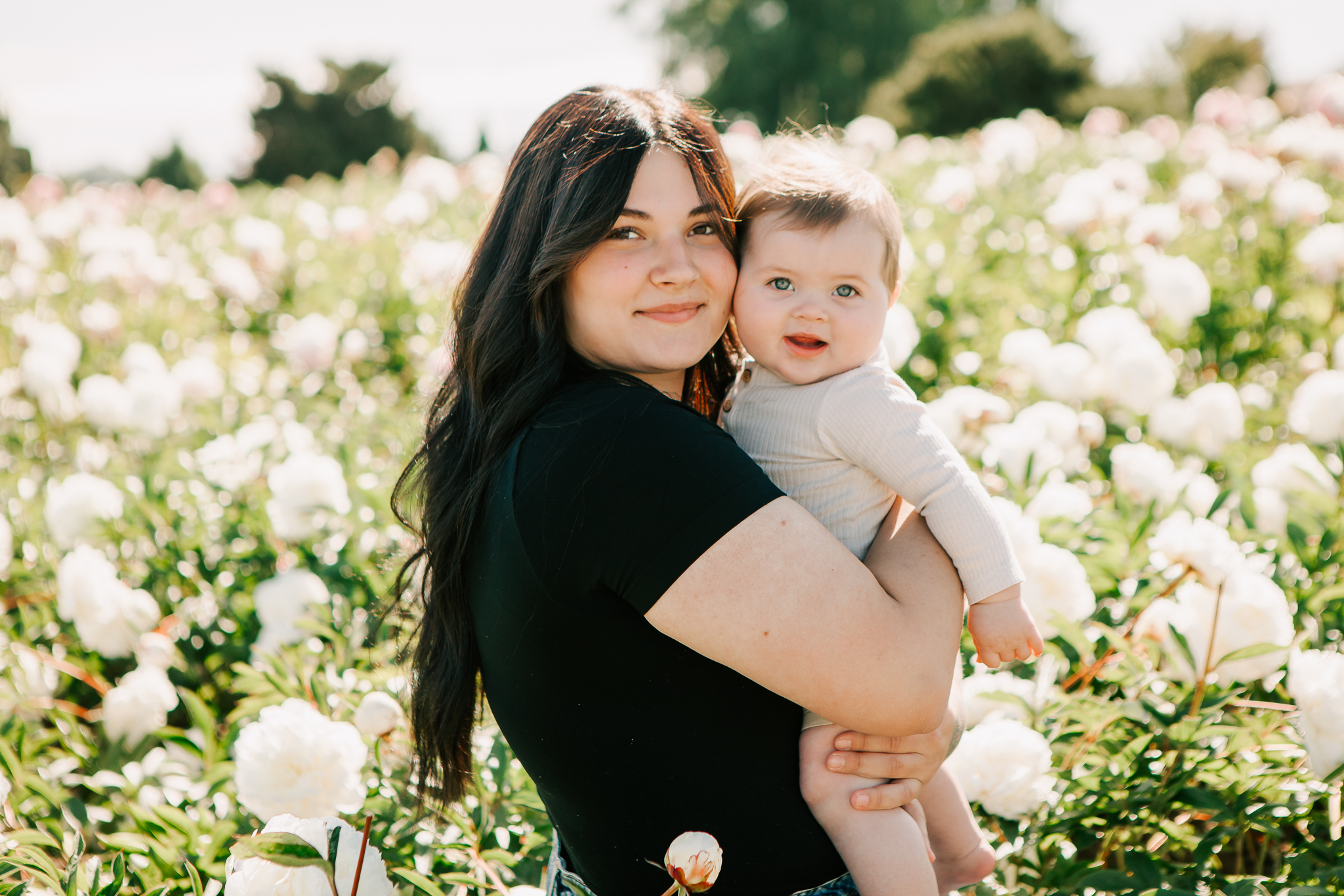 Mother holds her smiling baby in a field of flowers at a spring mini session at Adelman Peony Gardens in Salem, OR
