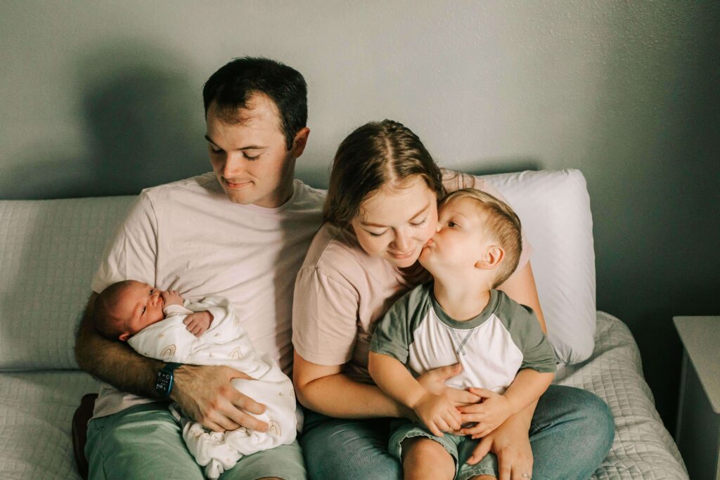 A family holds their newborn baby while sitting on a bed. Mom, dad, big brother, baby