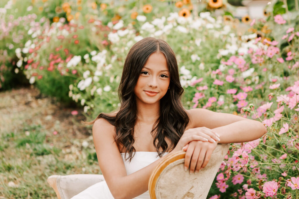 Senior girl in a flower patch at Rose Scott Farms, Albany, OR. She's sitting in a white dress on a cream chair with pink, white, and orange flowers.