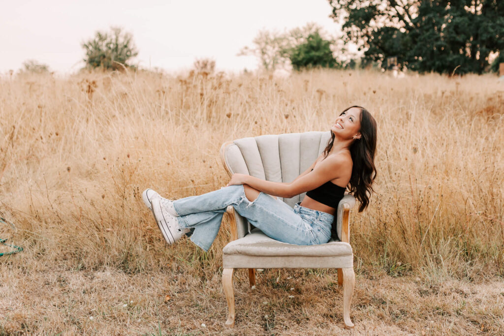 A teen girl poses in a chair for senior photos in a field in Albany, Oregon. She is wearing jeans and a black top and has her head thrown back laughing.