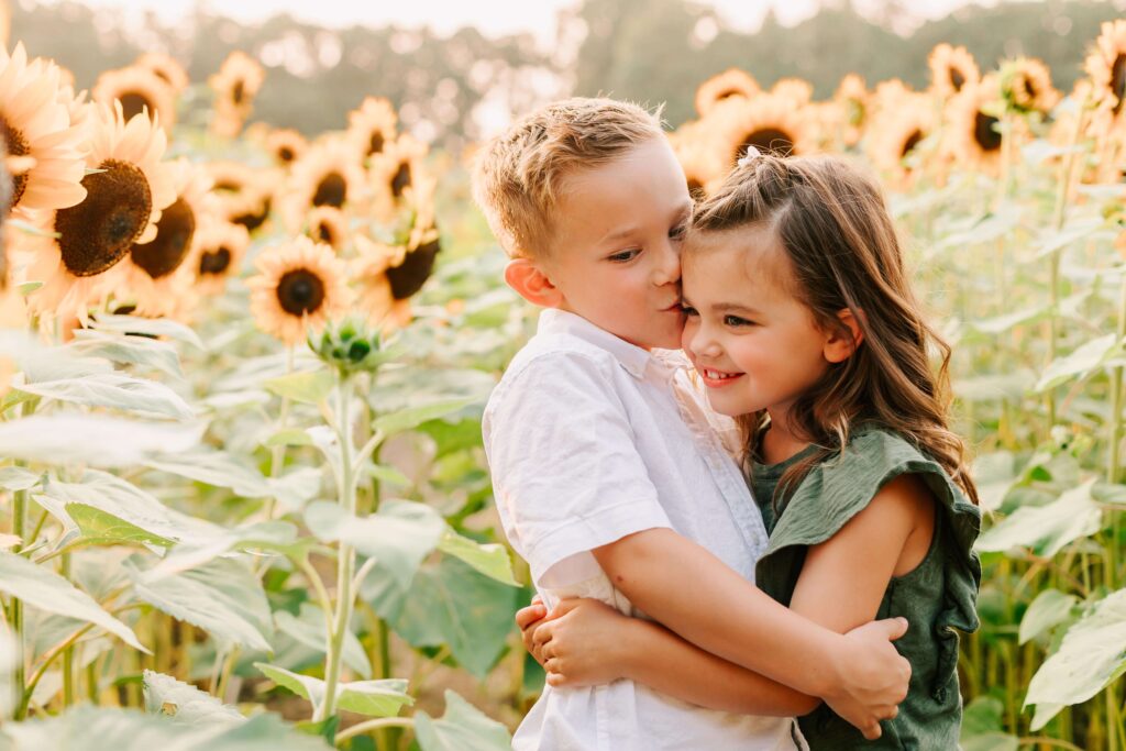 A young boy kisses his sister on the forehead. They're in a sunflower field in Albany.