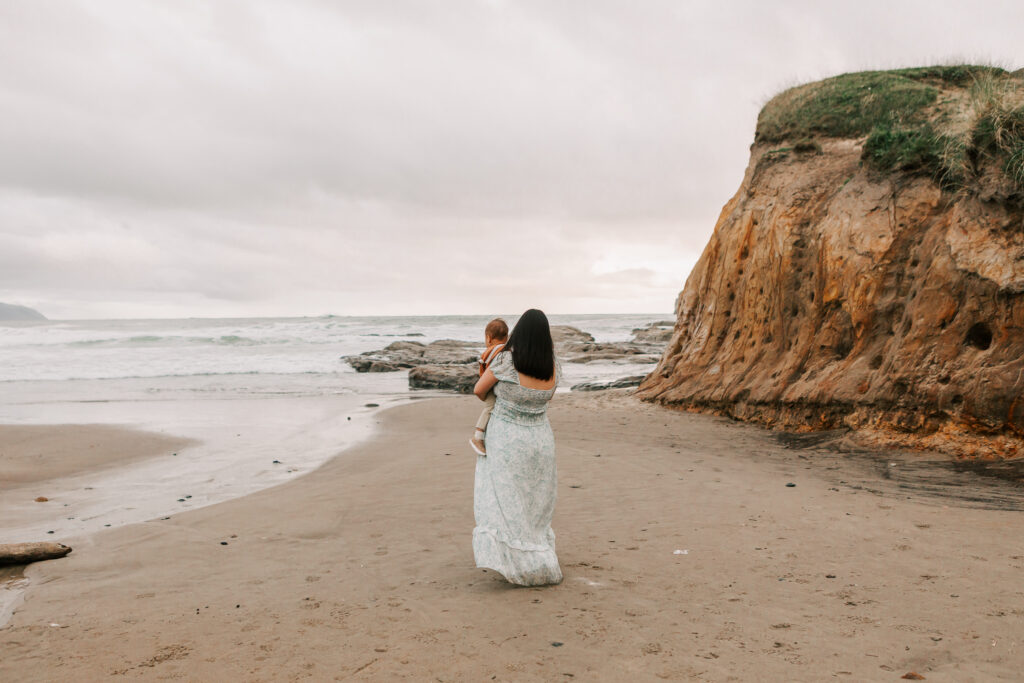 A mom holds her toddler and looks out at the ocean during a family photo session with natural posing with Jen White Photography 