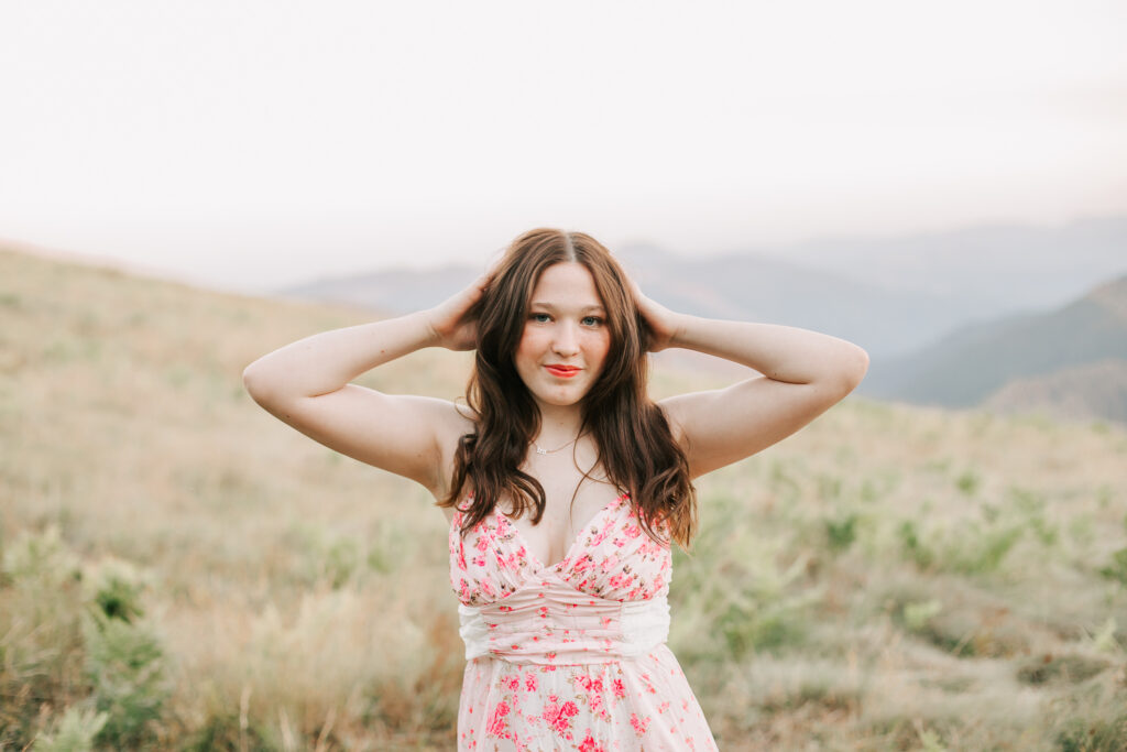 High school senior in a flowy dress posing in a sunlit field with mountains in the background, smiling at the camera