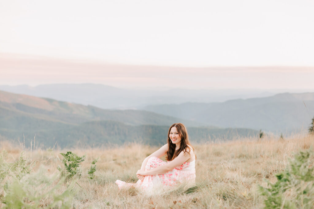 Senior portrait in flowy dress at Mary's Peak Philomath Oregon. She's sitting smiling at the camera with a sunset view in the background. Photo taken at a senior photo session by Jen White Photography from Albany Oregon