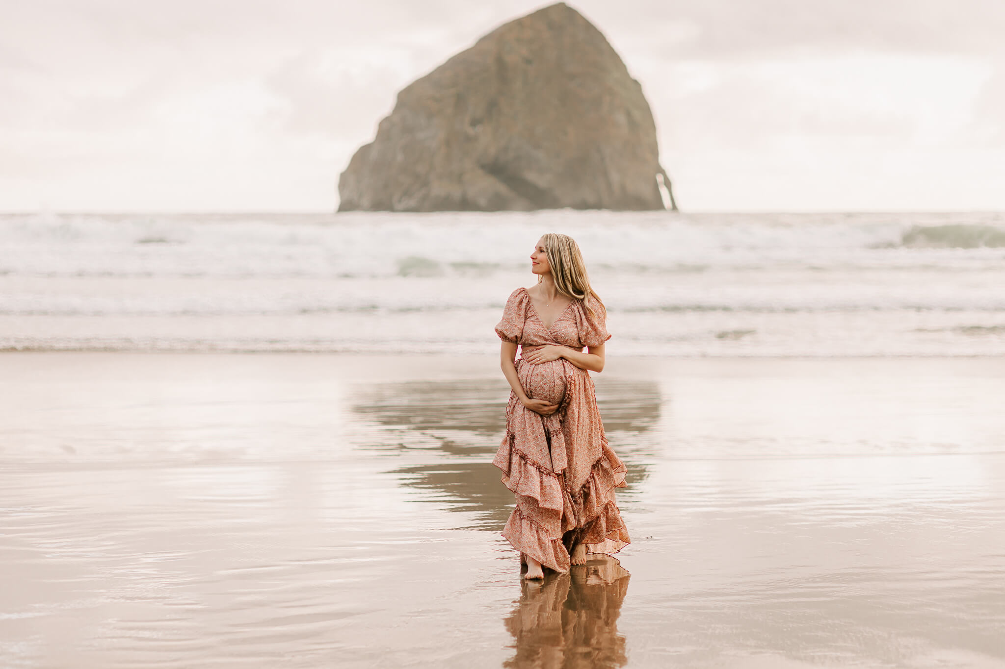 A pregnant mom stands at Cape Kiwanda in Oregon posing for maternity photos in a flowing floral gown