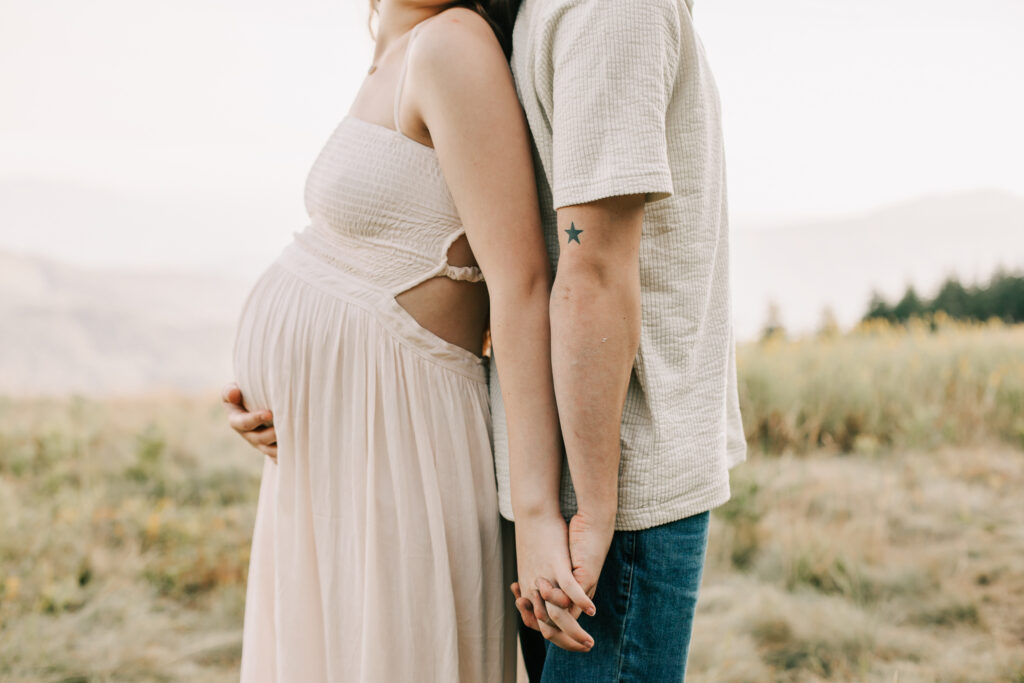 Man and woman stand back to back holding hands while posing for maternity photos. He has a small star tattoo on his upper arm