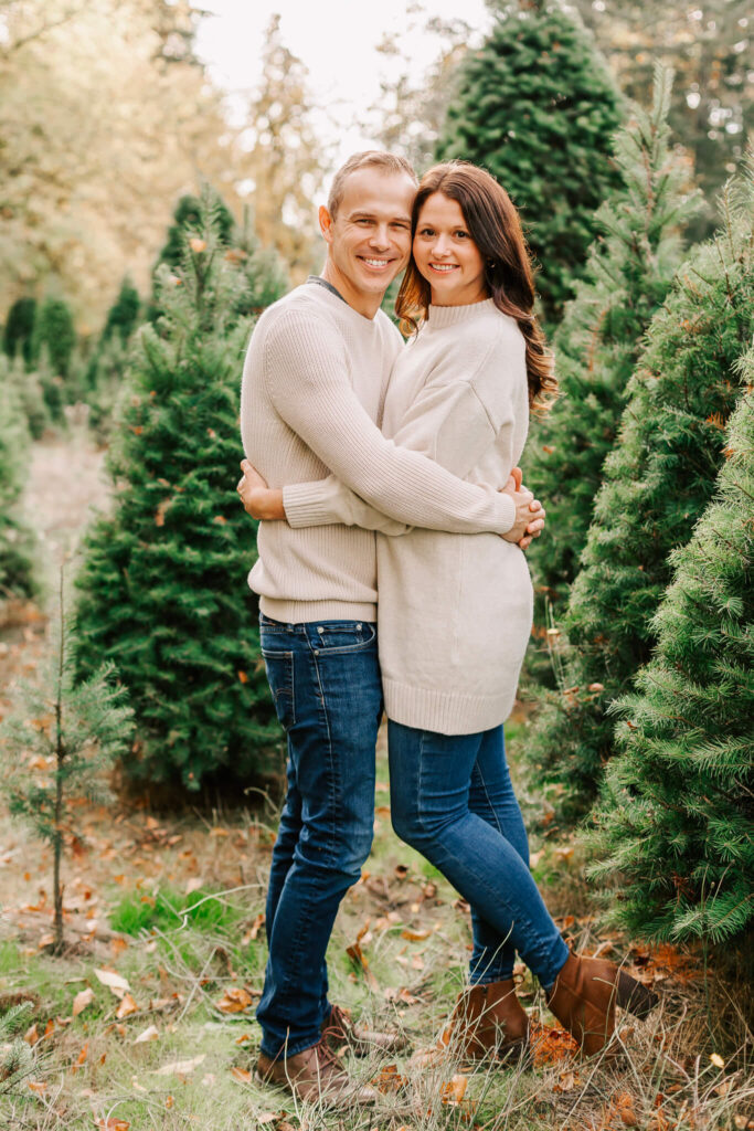 Husband and wife hug each other while looking at the camera. They're wearing sweaters and Jeans and posing at a Salem Oregon Christmas Tree farm mini event with Jen White photography