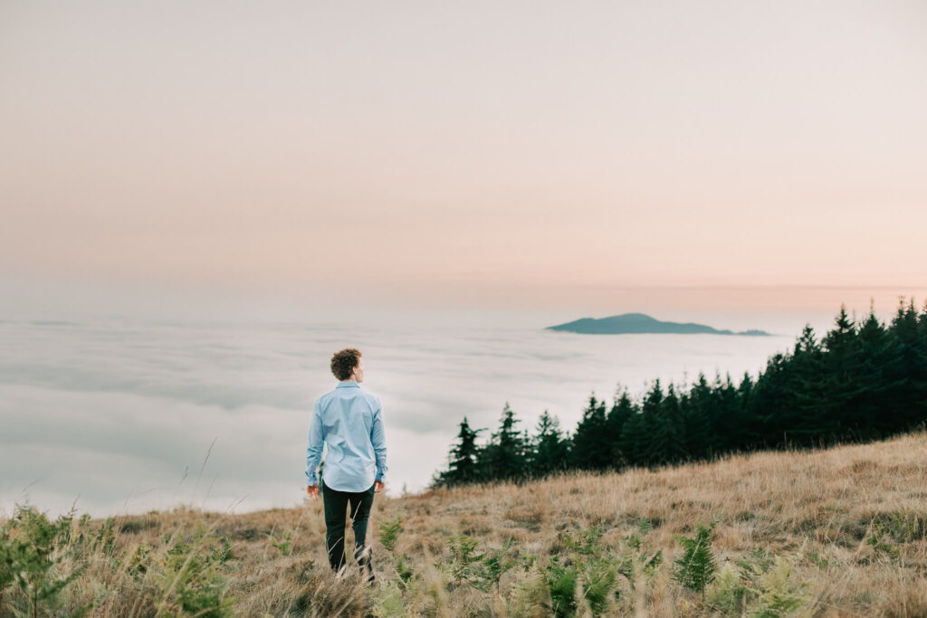 A senior boy walks in the mountains above the clouds during a session with Jen White Photography. He's wearing a blue shirt and black pants. He has curly hair and his back is to the camera. It's sunset over the clouds.