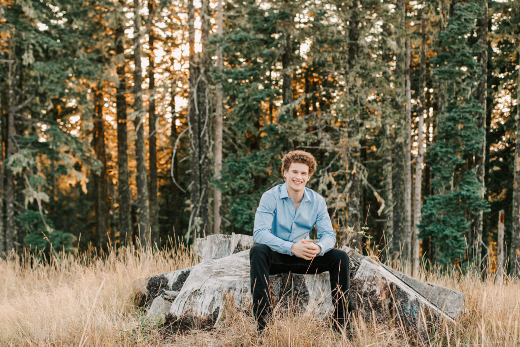 High school senior sits on a large stump with his arms resting on his knees in front of trees. He's wearing a blue shirt and black pants.
