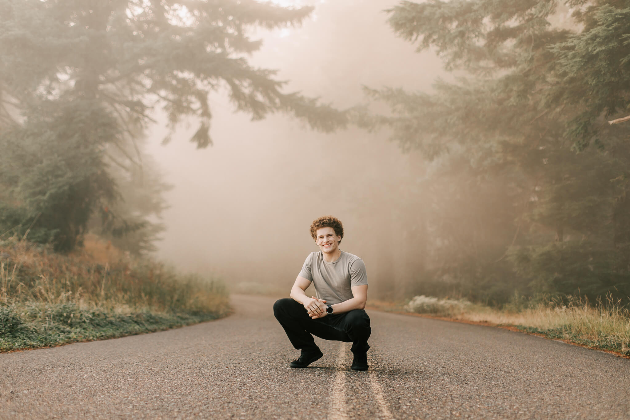 A high school senior squats in the road in the fog at Mary's Peak posing for a senior portrait with Jen White Photography of Albany Oregon. He's wearing a tan shirt and black pants.
