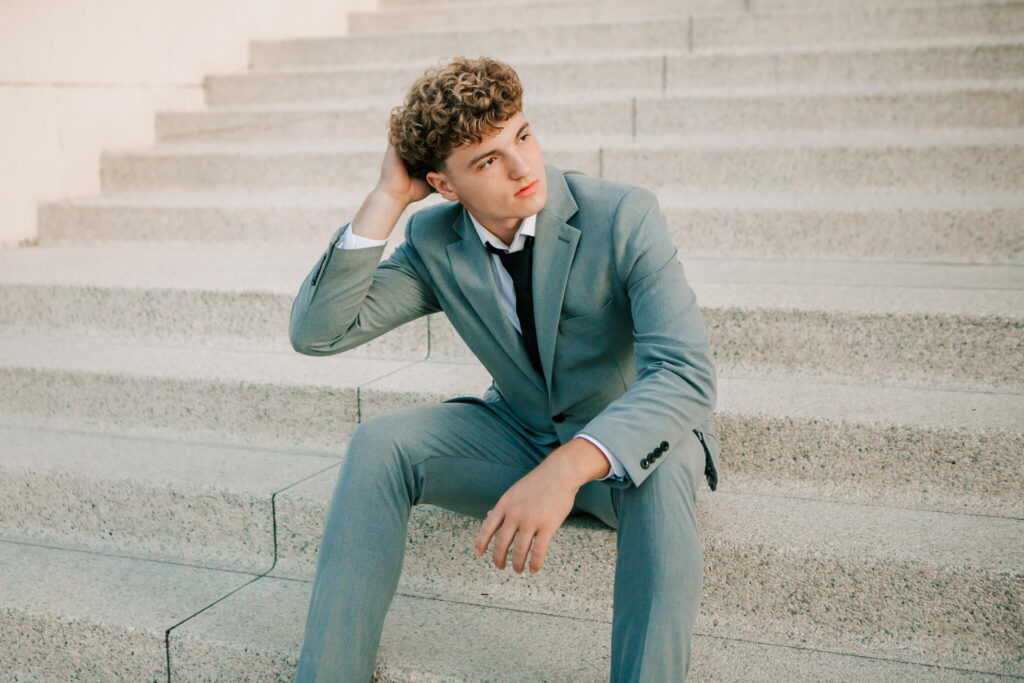 A senior boy in a gray suit sits on the steps looking to the side with his hand in his hair posing for senior portraits in Albany, OR