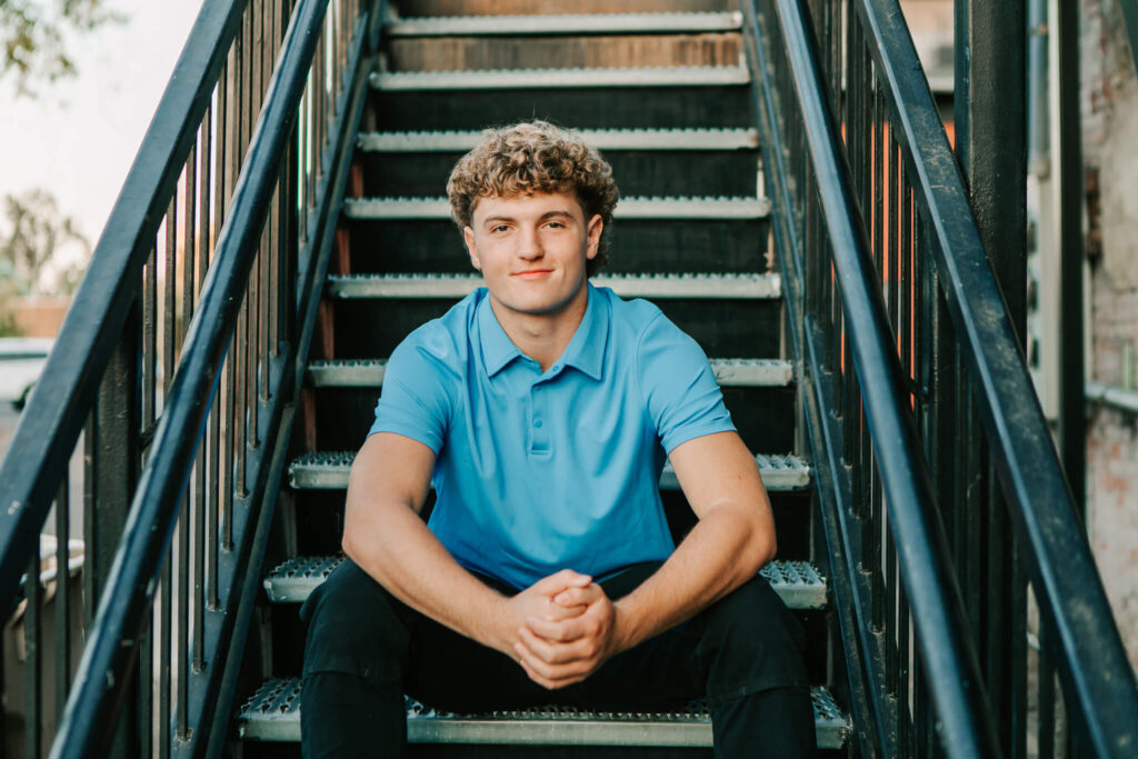 A high school senior in a blue polo sits on black steps posing for senior photos