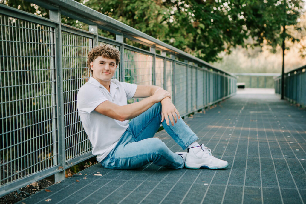 A senior boy sits on a bridge posing for senior portraits. He's wearing a white polo and blue jeans.