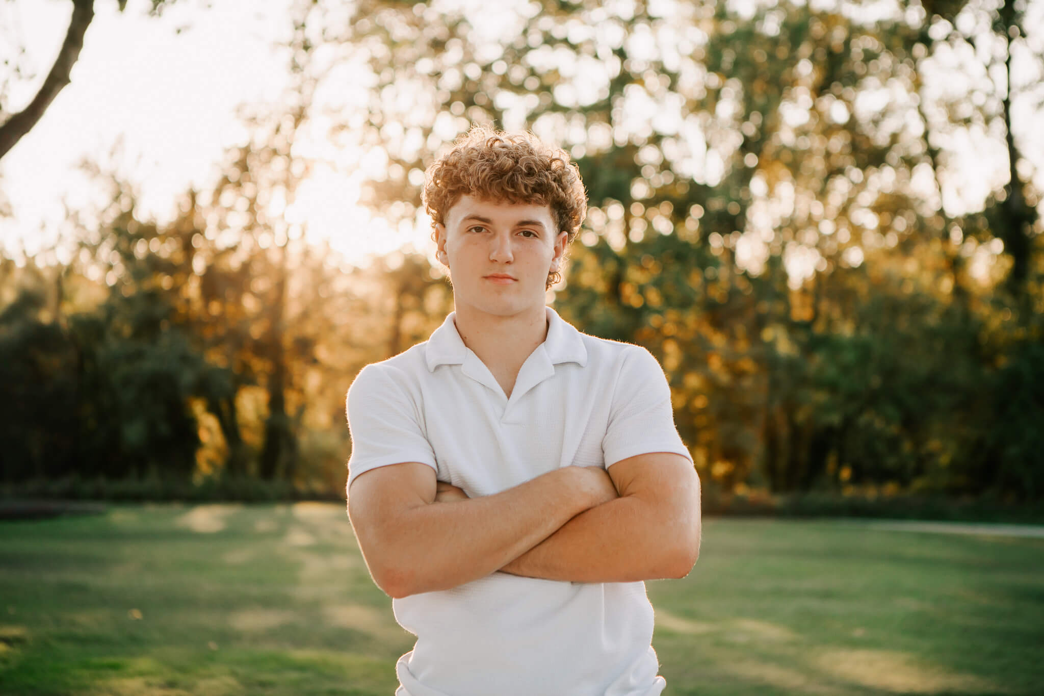 A high school senior stands in a field in a white polo with his arms crossed posing for senior portraits with Jen White Photography in Albany, Oregon