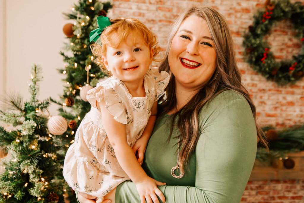 Mother and adorable red headed toddler girl smiling with a wreath and Christmas tree in the background during a mini session with Jen White Photography