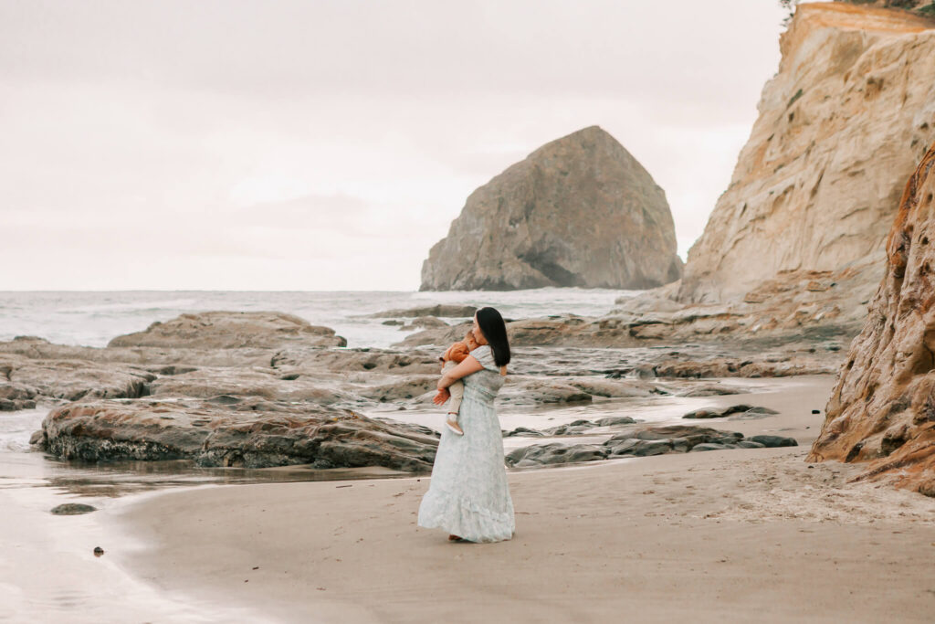 A mom holds her toddler son while standing in front of haystack rock on the Oregon coast