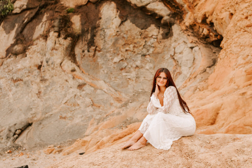 A young woman in a white dress sits at the bottom of a cliff on the oregon coast