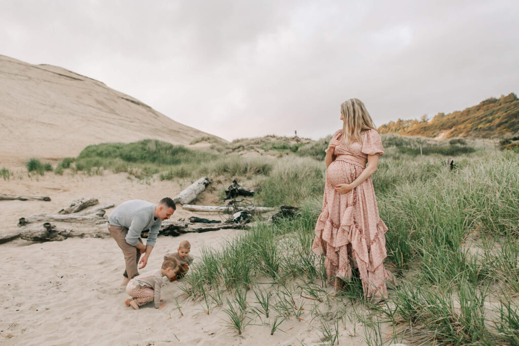 A pregnant mom watches her husband and toddler play on the beach in Oregon