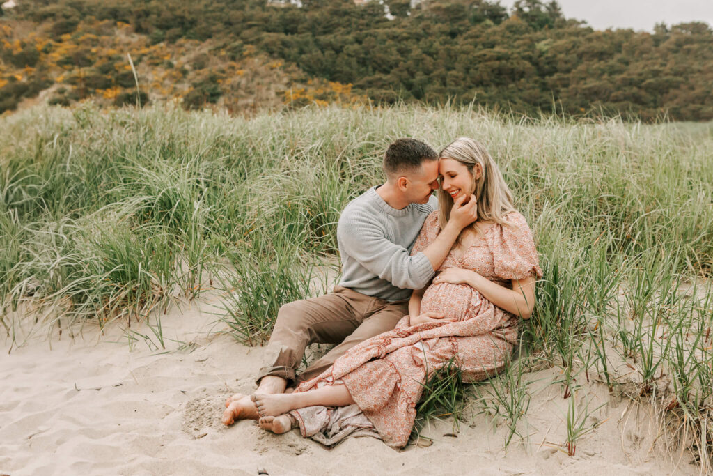 A husband and his pregnant wife sit in the beach grass at Cape Kiwanda State Park in Oregon