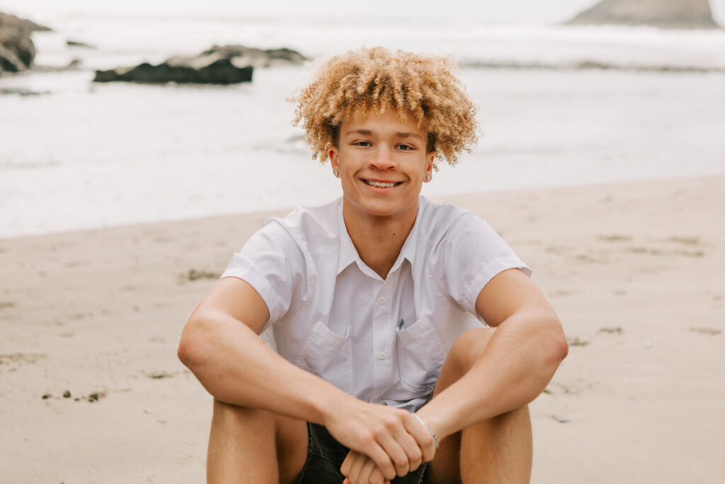 high school senior in a white shirt sits on the beach at Heceta Head in Florence 
