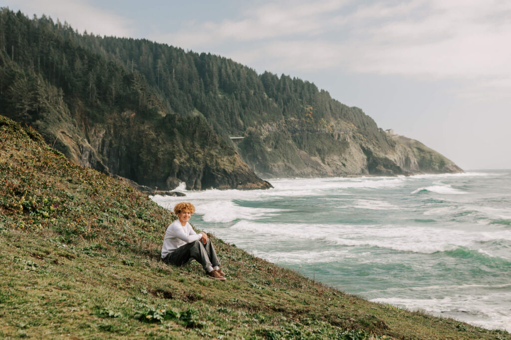 High school senior sits on a grassy cliff with trees in the background looking over the ocean at Heceta Head Lighthouse while posing for senior portraits with Jen White Photography of Albany Oregon
