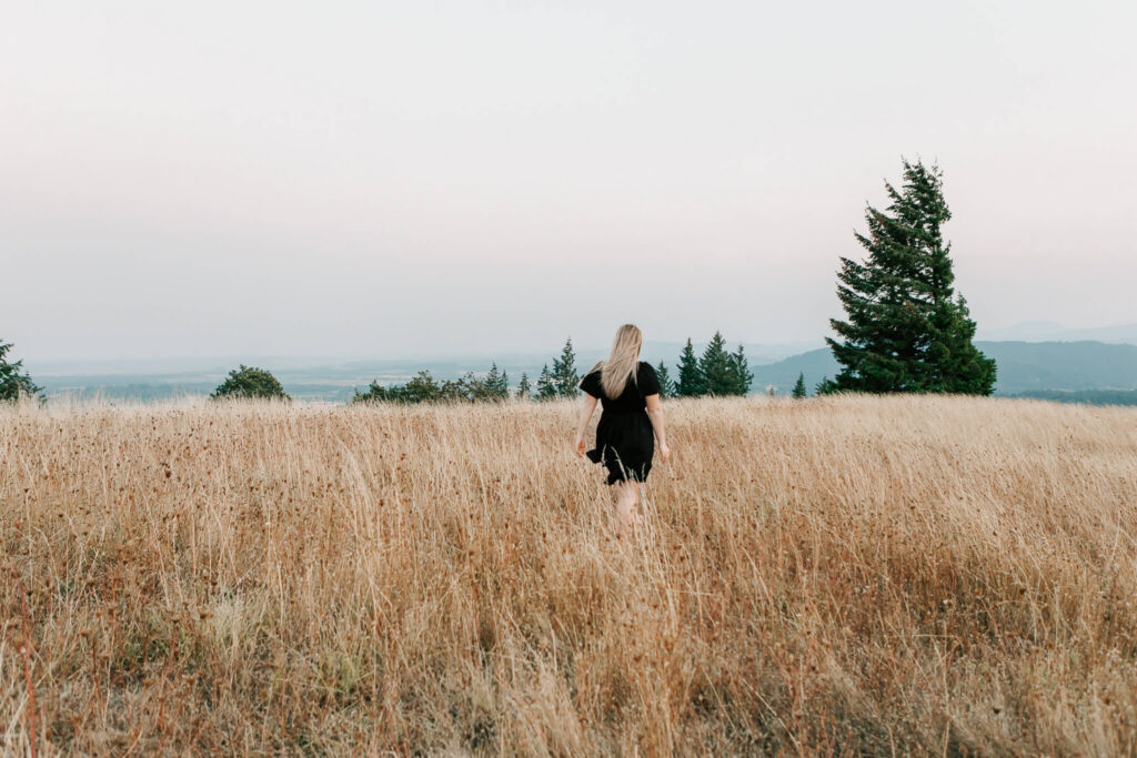 girl in black dress walking in golden field with trees in the background