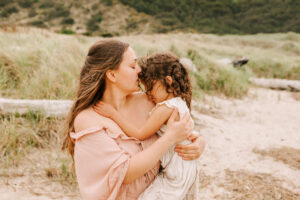Mom kisses her young daughter on the head. They're on the beach