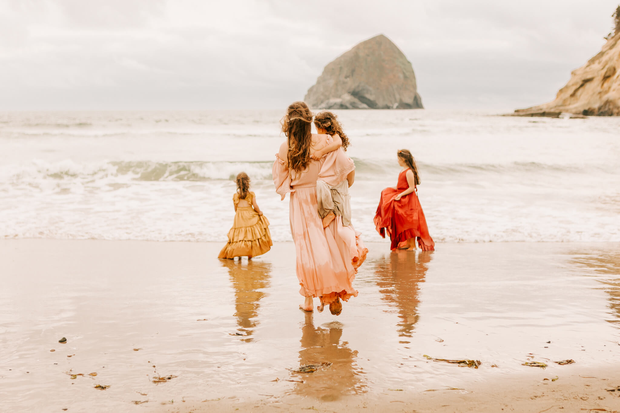 A mother holds her young daughter while walking in the ocean at Cape Kiwanda Oregon with her other 2 daughters